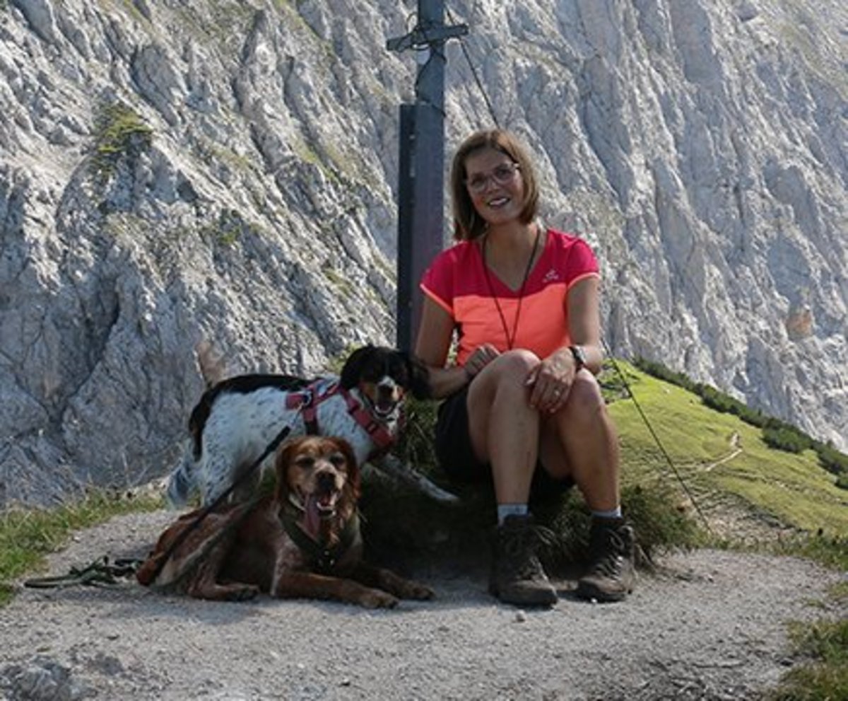 Carola sitzt mit ihren zwei Hunden vor einem Gipfelkreuz in den Tiroler Bergen. 