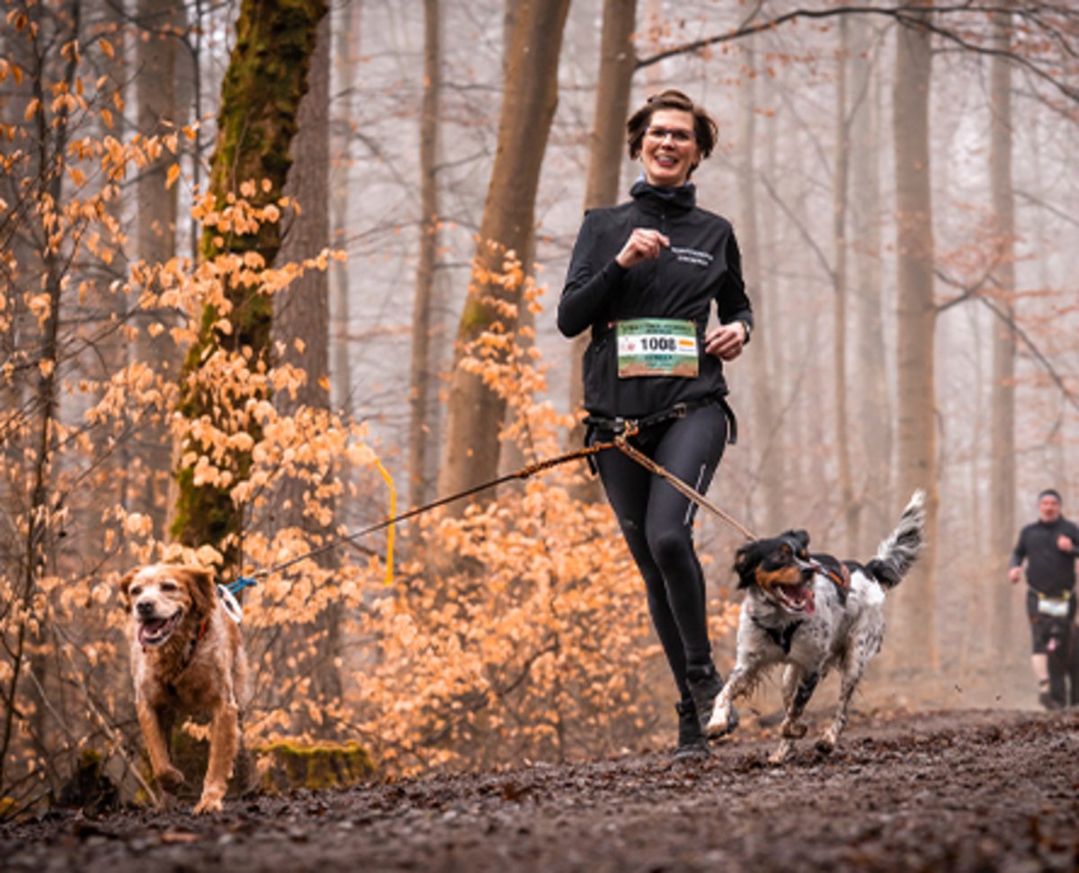 Carola joggt lachend mit ihren beiden Hunden durch einen herbstlichen Wald. 