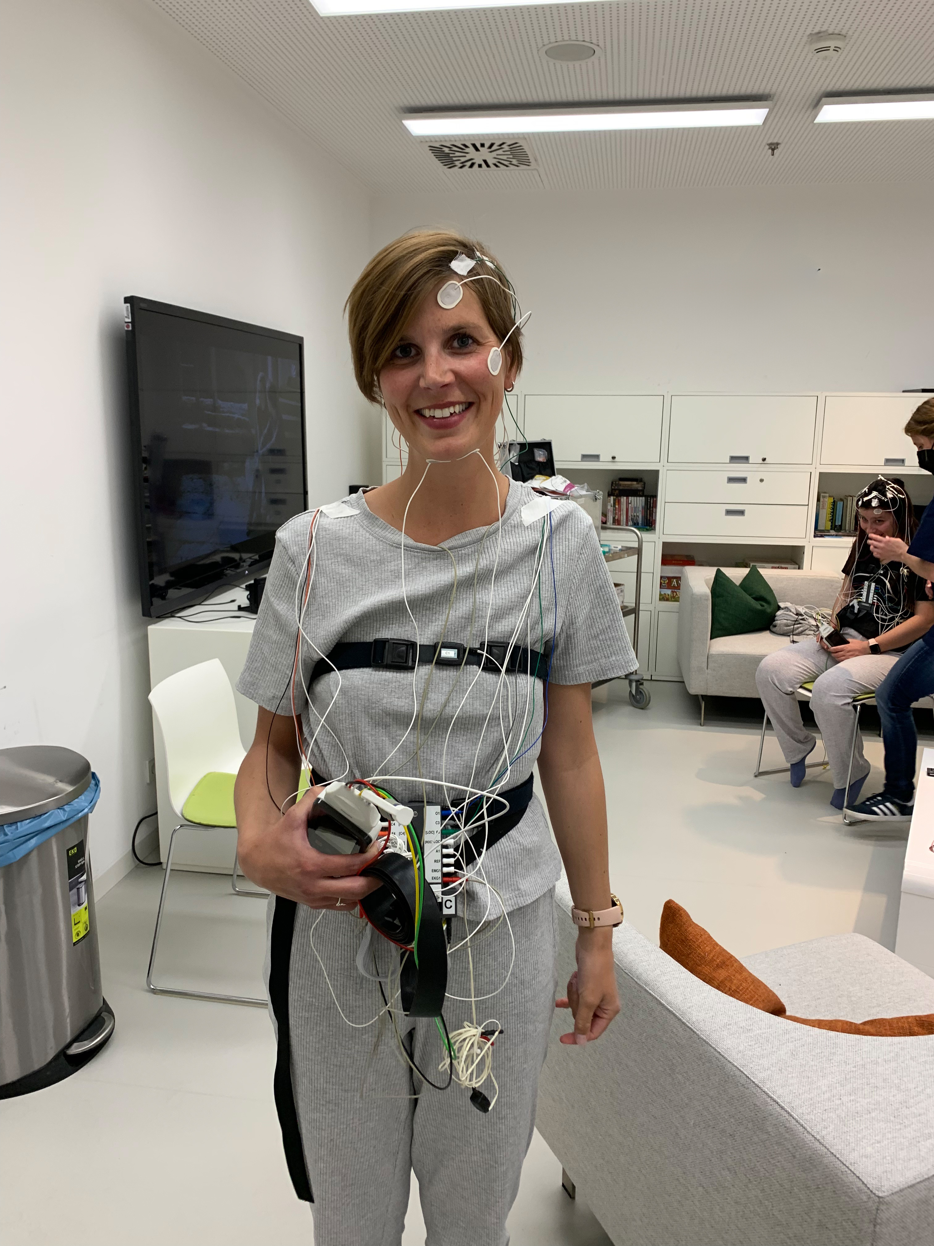 Carola steht lächelnd in einem Raum des Deutschen Zentrums für Luft- und Raumfahrt (DLR) und trägt zahlreiche Sensoren und Kabel am Kopf und Körper, für eine wissenschaftliche Untersuchung.